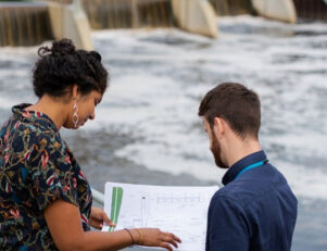 Uma mulher e um homem, analisando um projeto, com uma represa ao fundo