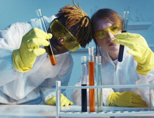 uma mulher jovem branca e um homem jovem negros os dois com oculos de proteção amarelo segurando um tubo de ensaio imagem utilizada para artigo sobre preço do curso de quimica