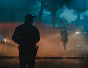 Homem parado em frente a uma faixa policial em uma cena de crime durante a noite